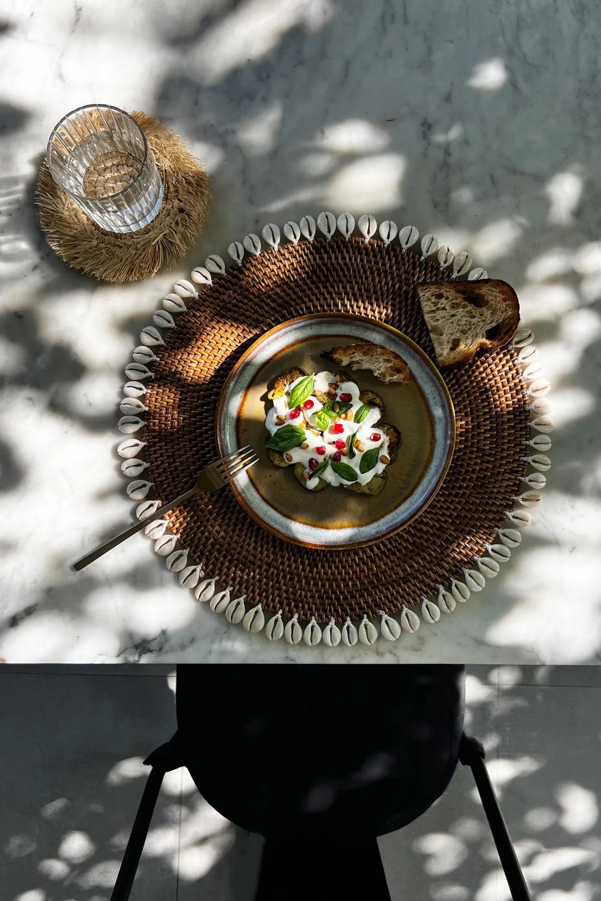 Un set de table en rotin et cauris dressé sur une table en marbre, avec une assiette garnie, du pain et un verre