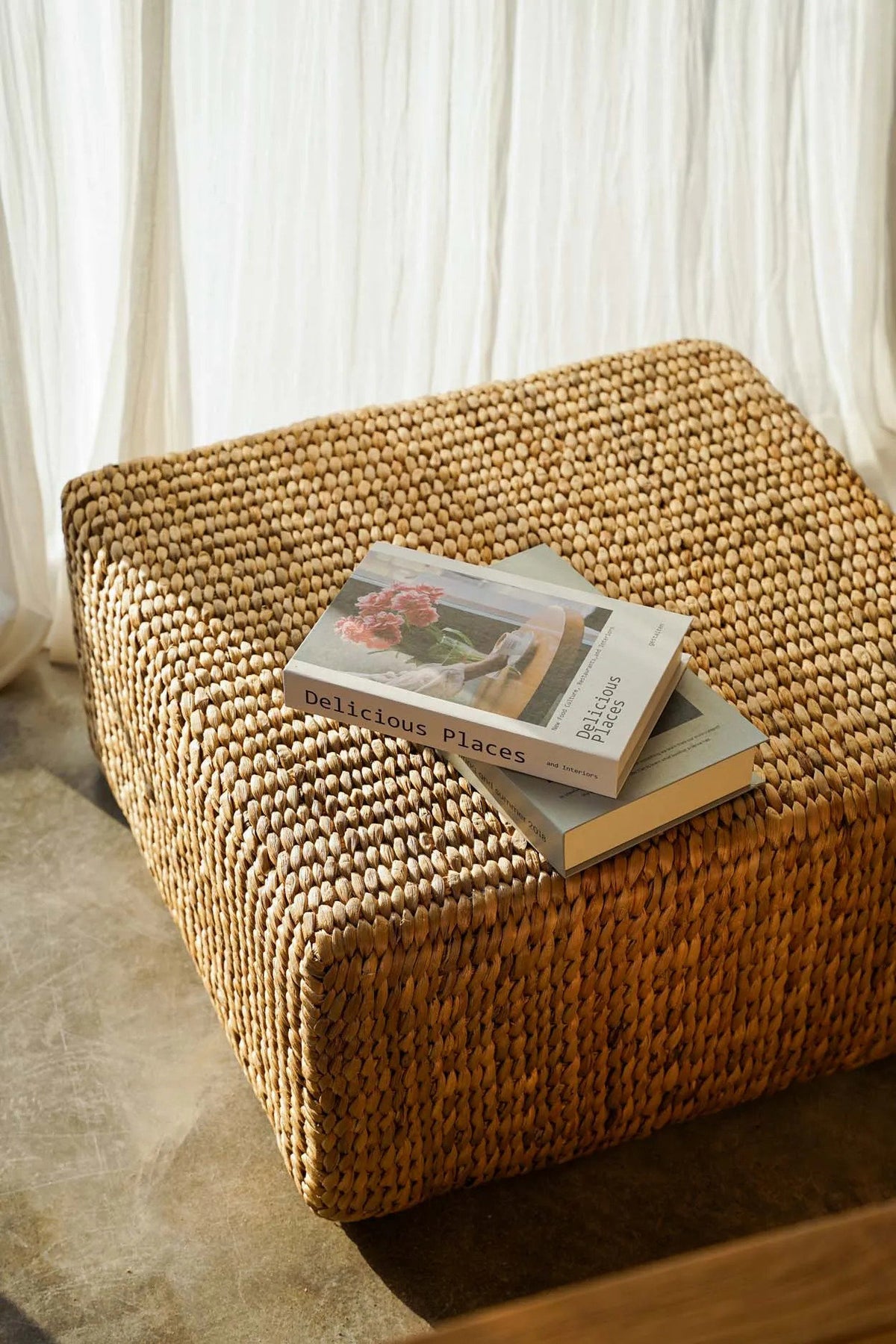 Pouf carré en jacinthe d'eau devant une banquette en teck, livres posés dessus