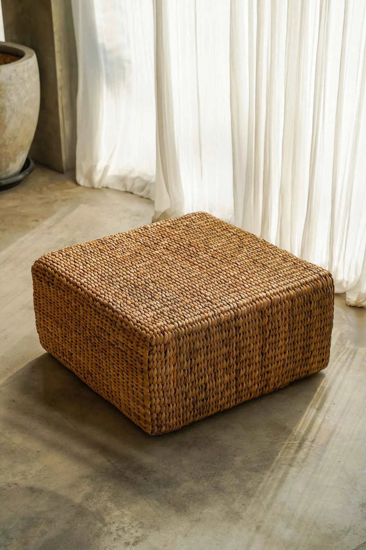 Pouf carré en jacinthe d'eau dans un intérieur clair, à côté d'une jarre en terre cuite